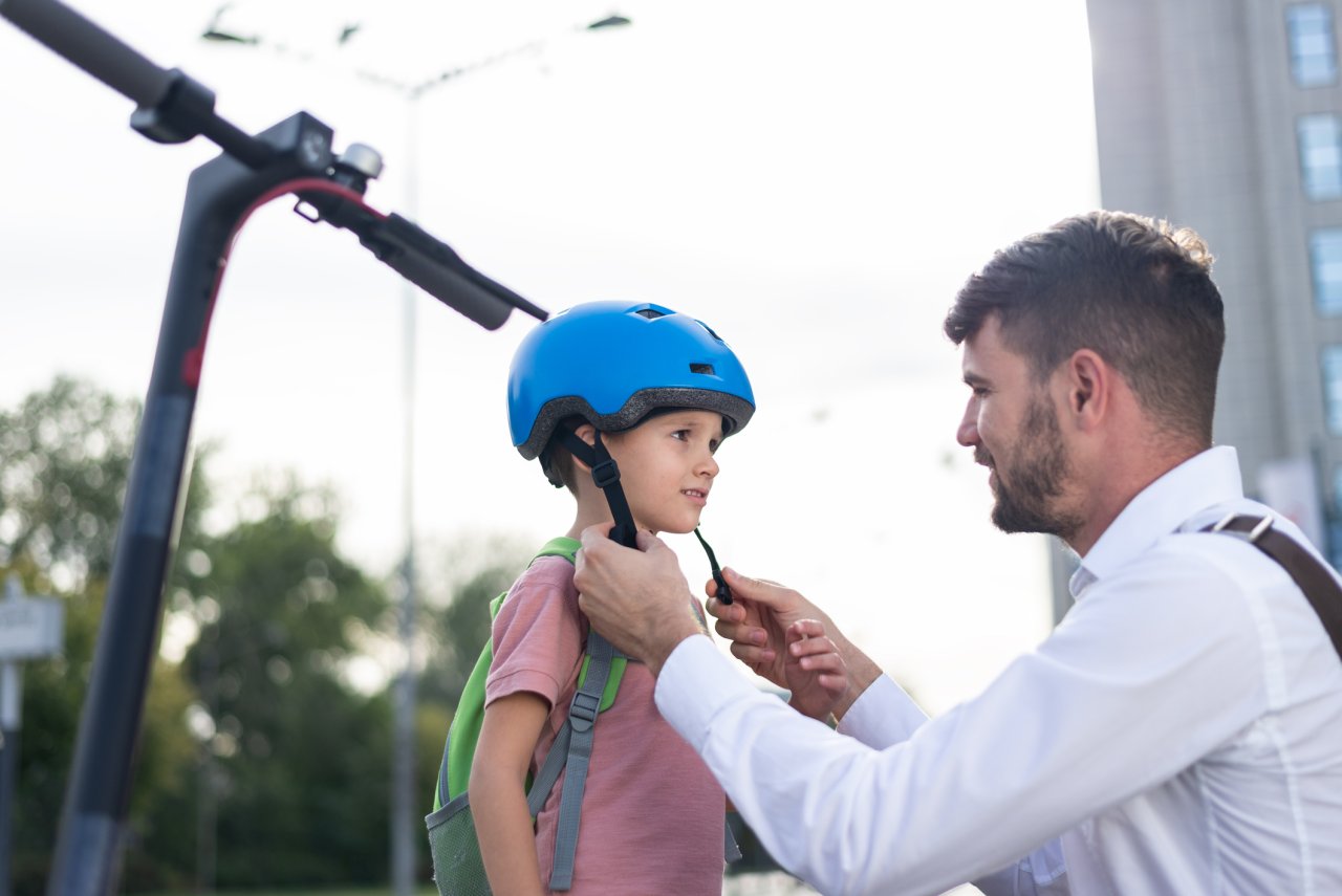 Ein Vater setzt seinem Sohn einen Schutzhelm für das E-Scooter-Fahren auf und verschließt gerade die Kinnriemen. 