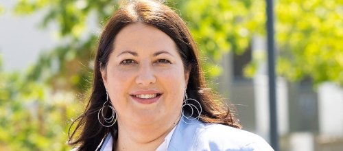 Im Vordergrund ist Frau Kathrin Gaál zu sehen. Sie steht und lächelt in die Kamera. Im Hintergrund sieht man zwar urbane Umgebung, aber mit viel Grün.