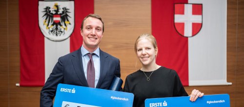 Die Preisträger*innen Moritz Muckenhuber und Anna Plötzl halten ihren Siegerscheck in der Hand und lächeln in die Kamera. Im Hintergrund sind zwei Flaggen (Österreich und Wien) zu sehen, die an der Wand hängen.