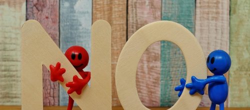 Auf dem Foto ist in hölzernen Buchstaben das Wort "No", also Nein, zu sehen, die von jeweils einer Spielzeugfigur, eine in blau, die andere in rot, gehalten werden.