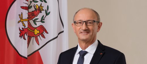 Der Tiroler Landeshauptmann Anton Mattle, steht im dunklen Anzug und mit Krawatte vor der Tiroler Fahne.