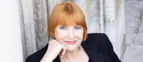 Auf dem Bild ist Chris Lohner zu sehen. Sie sitzt und blickt direkt in die Kamera. Ihre rechte Hand stützt ihre rechte Wange. Ihr Ausdruck wirkt freundlich und aufmerksam. Der Hintergrund ist neutral und lenkt nicht von der Person ab.