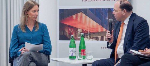 Auf dem Podium befinden sich die Moderatorin und Ehrenpräsident der Kammer für Ärztinnen und Ärzte in Wien, Thomas Szekeres, der gerade spricht und ein Mikrofon in der rechten Hand hält. Beide sitzen, zwischen ihnen steht ein kleines Tischchen, auf dem sich zwei Mineralwasserflaschen befinden sowie zwei Gläser. Im Hintergrund ist ein Plakat der Veranstaltung zu erkennen.