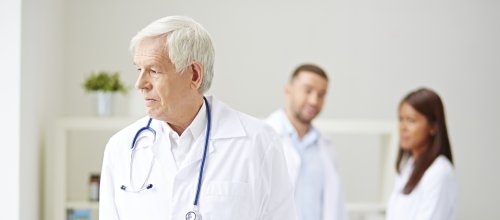 Ein älterer Arzt blickt nachdenklich zur Seite. Er hat einen weißen Arztkittel an und trägt ein Stehtoskop um den Nacken. Hinter ihm stehen, in einer Unschärfe zwei Personen - ein Mann und eine Frau.