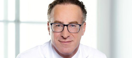 Der Arzt Shahrokh Shariat sitzt an einem Tisch, die Hände vor sich zusammengetan, und hat einen weißen Arztkittel an und trägt eine blaue Krawatte. Er trägt ebenso eine Brille und blickt direkt in die Kamera.