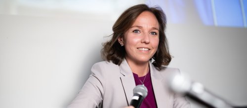 Ein Frau sitzt in einem Fauteuil, hält ein Mikrofon in der Hand und lächelt in die Kamera.