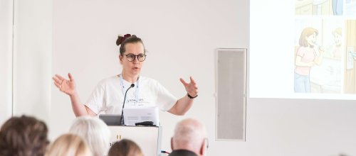 Eine Frau hält gerade einen Vortrag, spricht und hält beide Hände und Arme vor sich, so als würde sie ein Längenmaß beschreiben.