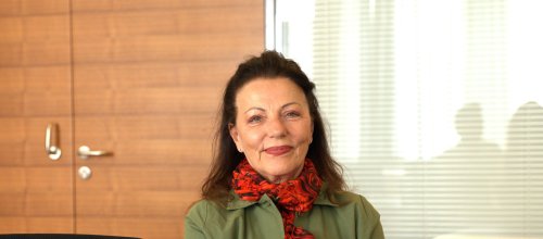 Eine Frau mit rotem Schal sitzt an einem Tisch mit nach vorne verschränkten Armen und blickt direkt in die Kamera.