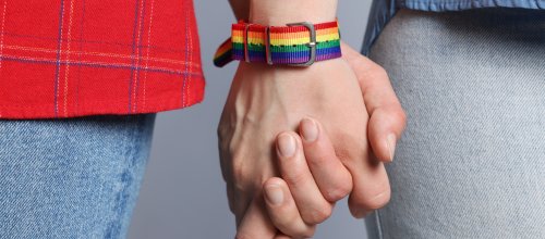 Zwei Hände mit einem Regenbogenarmband halten einander fest.