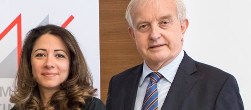 Zwei Personen, eine Frau, die links steht, und ein Mann, der rechts steht, blicken freundlich und direkt in die Kamera.