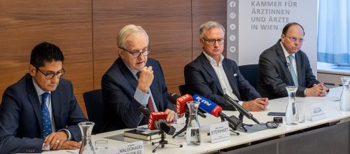 Vier Männer sitzen auf einem Tisch im Rahmen einer Pressekonferenz.