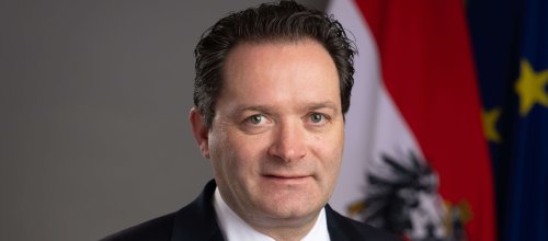 Der Bundesminister für Landwirtschaft Norbert Totschnig blickt in die Kamera - hinter ihm sieht man Teile der Fahnen von Österreich und der EU.