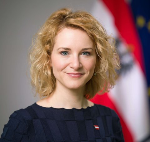 Die Bundesministerin Eva-Maria Holzleitner blickt in die Kamera, im Hintergrund sieht man die Österreichische Fahne, sowie einen kleinen Teil der EU-Flagge