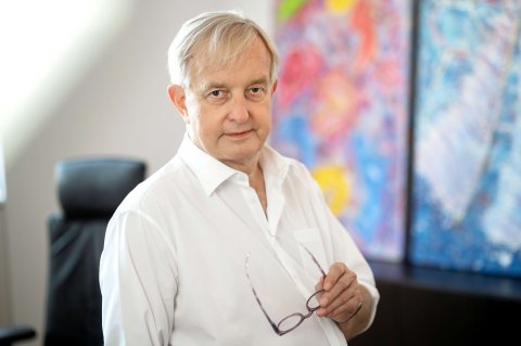 Präsident Johannes Steinhart in seinem Büro, hält seine Brille in der linken Hand und blickt in die Kamera. Im Hintergrund sind Bilder zu sehen.