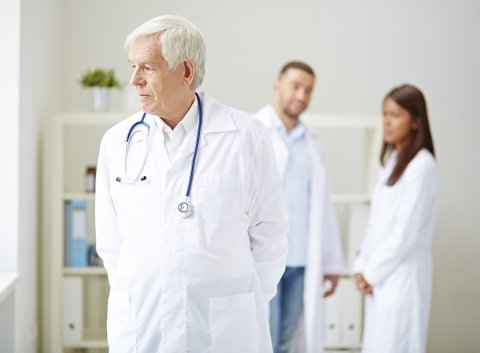 Ein älterer Arzt blickt nachdenklich zur Seite. Er hat einen weißen Arztkittel an und trägt ein Stehtoskop um den Nacken. Hinter ihm stehen, in einer Unschärfe zwei Personen - ein Mann und eine Frau.