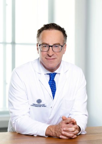 Der Arzt Shahrokh Shariat sitzt an einem Tisch, die Hände vor sich zusammengetan, und hat einen weißen Arztkittel an und trägt eine blaue Krawatte. Er trägt ebenso eine Brille und blickt direkt in die Kamera.