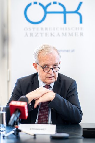 Mann mit Brille sitzt an einem Tisch mit Mikrofonen.