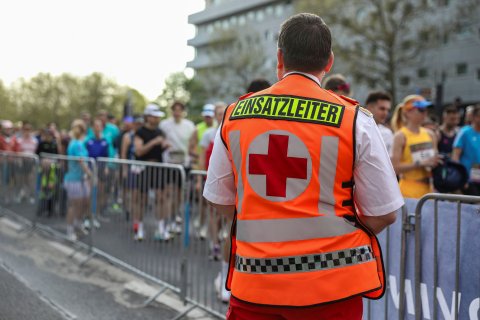 Der Einsatzleiter vom Roten Kreuz steht mit dem Rücken zur Kamera und blickt auf die Läuferinnen und Läufer, die man hinter einer Absperrung auf einer Straße laufen sieht.