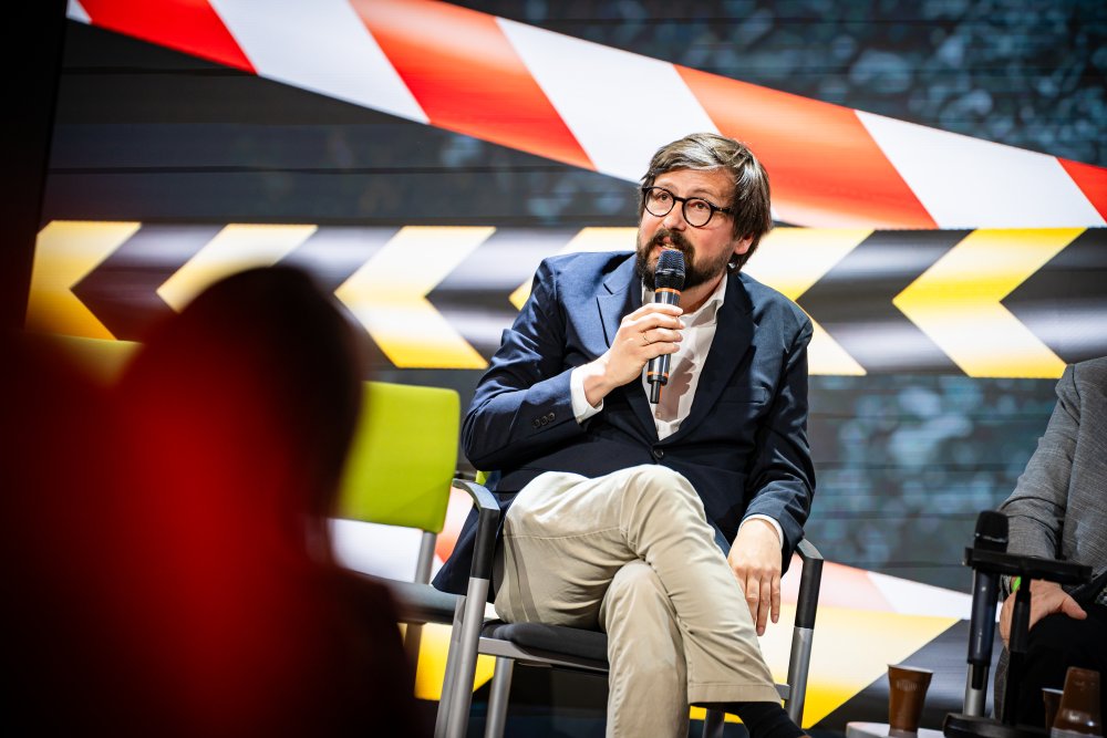 Dunkelhaaringer Mann mit Brille sitzt auf einer Bühne und hält ein Mikrofon.