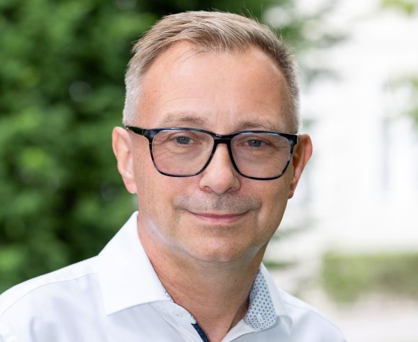 Ein Porträt des Arbeits- und Schmerzmediziners Richard Crevenna. Aufgrund des Hintergrunds kann geschlossen werden, dass die Aufnahme im Außenbereich stattgefunden hat - verschwommen ist ein Baum oder eine Hecke im Hintergrund zu sehen. 