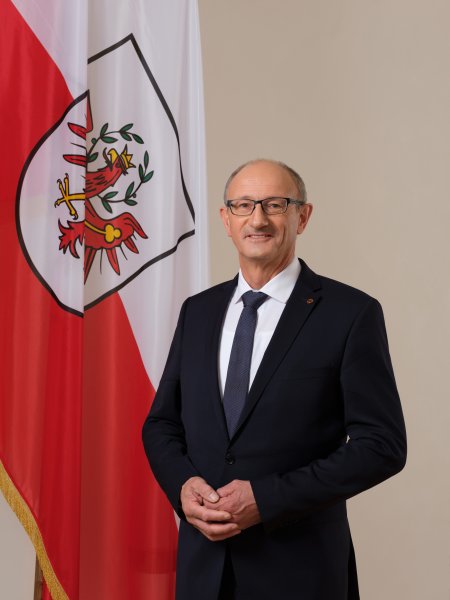Der Tiroler Landeshauptmann Anton Mattle, steht im dunklen Anzug und mit Krawatte vor der Tiroler Fahne.