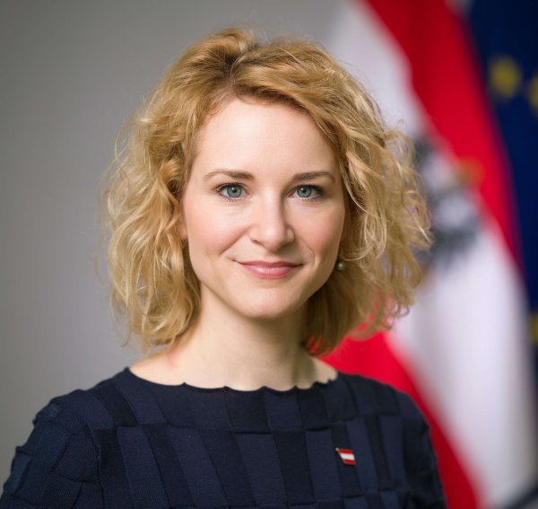 Die Bundesministerin Eva-Maria Holzleitner blickt in die Kamera, im Hintergrund sieht man die Österreichische Fahne, sowie einen kleinen Teil der EU-Flagge