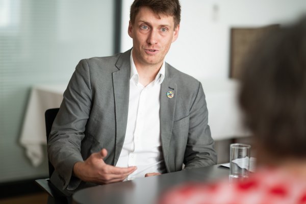 Mann mit Hemd und Sakko an einem Tisch sitzend, zum Interviewpartner sprechend.