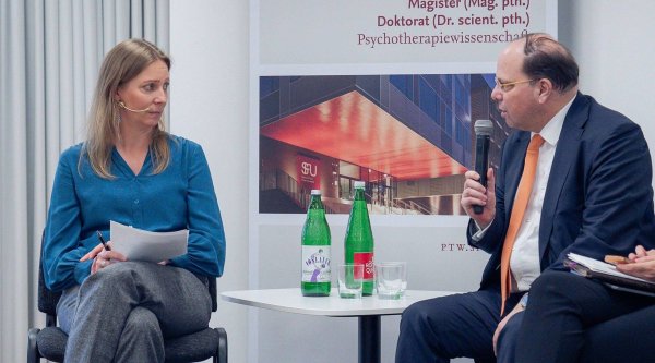 Auf dem Podium befinden sich die Moderatorin und Ehrenpräsident der Kammer für Ärztinnen und Ärzte in Wien, Thomas Szekeres, der gerade spricht und ein Mikrofon in der rechten Hand hält. Beide sitzen, zwischen ihnen steht ein kleines Tischchen, auf dem sich zwei Mineralwasserflaschen befinden sowie zwei Gläser. Im Hintergrund ist ein Plakat der Veranstaltung zu erkennen.
