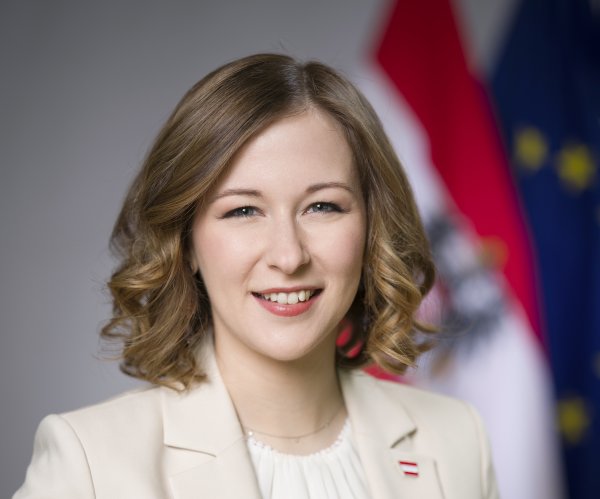 Frau mit Sakko vor österreichischer Fahne und EU-Fahne lächelt in die Kamera.