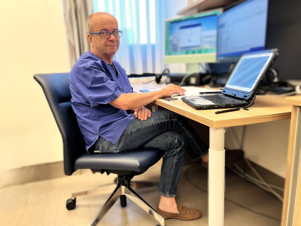Ein Mann sitzt in einem Sessel vor einem Schreibtisch, auf dem ein Laptop und zwei zusätzliche Bildschirme stehen.